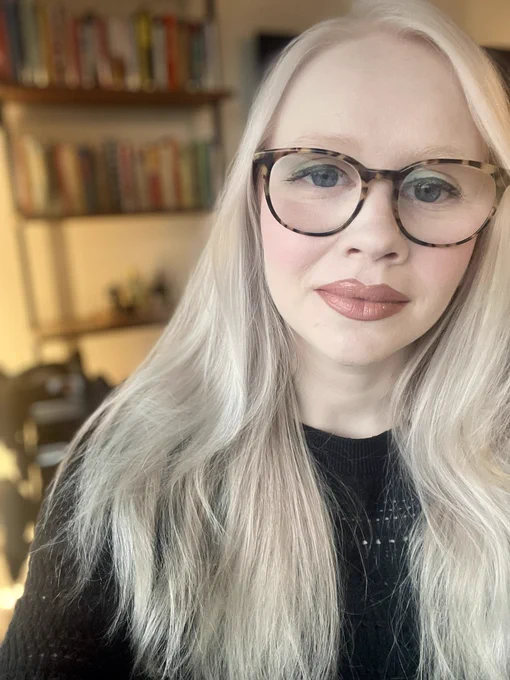 A picture of me in a black sweater and glasses looking at the camera. My hair is down and there’s a bookshelf in the background.
