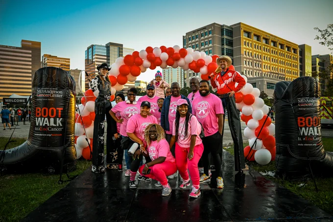 A group of people in pink t-shirts and two people on stilts posing in front of a balloon arch