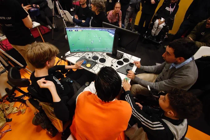 4 personnes vues de haut, jouant à un jeu de foot, autour d'une même console et d'un meme écran 
