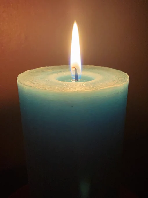 A large, newly lit turquoise candle against a rusty brown background. On the lower half of the picture the candle disappears into darkness.