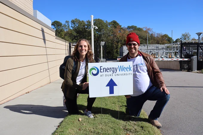 Two people crouched behind a sign for Energy Week at Duke University outside a chiller plant.