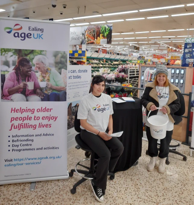 Image shows two female volunteers in Age UK Ealing t-shirts sitting in Tescos next to Age UK Ealing signage, with a collection bucket and membership leaflets.