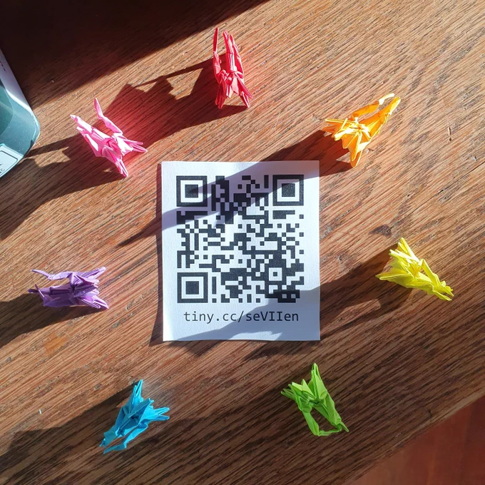 Seven origami dragons, each a different colour form a rainbow ring around a QR code lying on a bookshelf in the sun.