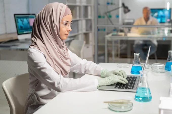 Woman in hijab and white coat with safety glasses and gloves in lab typing on laptop with conical flasks and petri dishes around her on desk.