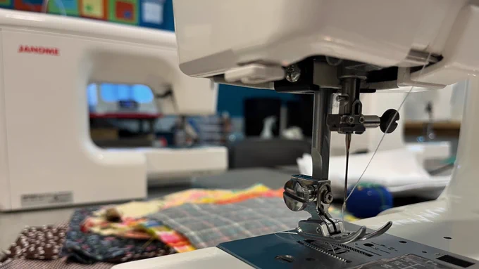 Closeup of a sewing machine needle with another machine and fabric pieces in the background