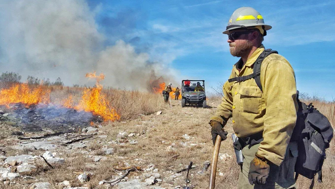 A firefighter stands close to the camera looking away while other firefighters work in the background with a UTV next to a prescribed fire.