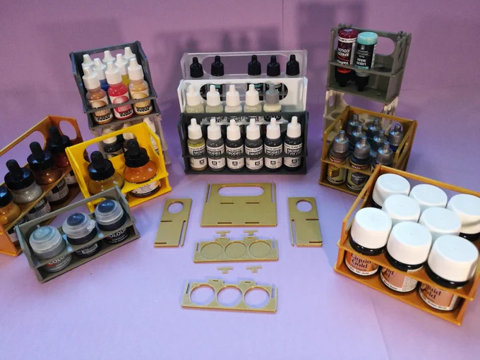 Collection of several paint bottle stands, holding bottles and pots of different brands: Liquitex Ink, Citadel paints, Daler Rowney ink, Jacquard Pinata, Vallejo Model Color and Game Color, Army Painter and Vallejo Liquid Gold. Some of the stands are stacked. There are three with Vallejo paints stacked in a three-step ladder. Some of the stands have several rows. A dismounted stand is also displayed, showing an exploded view with all its parts: two side panels, a back panel, a top tray with full holes for the bottles, a bottom tray with blind holes for the bottles, and tray locking pins.