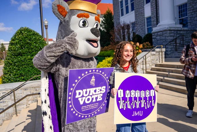 Duke Dog poses for a photo with a student holding an “I Voted” sign. Duke Dog holds a “Dukes Vote” sign.
