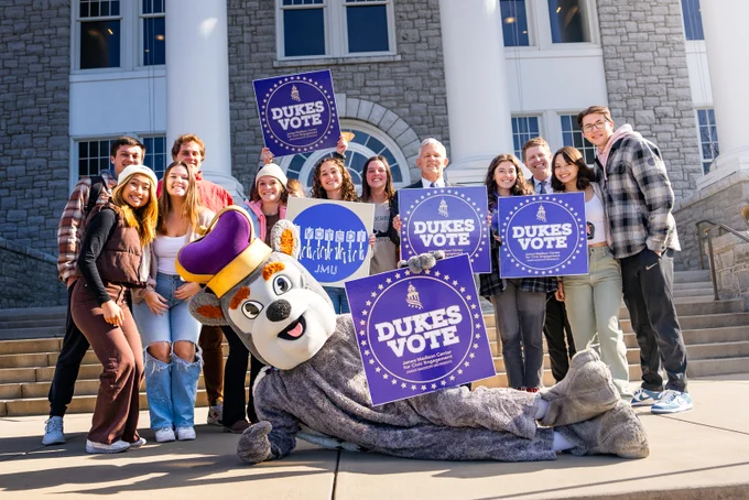 Duke Dog lays on their side holding a “Dukes Vote” sign. Behind him is a group of students and President Alger also holding “Dukes Vote” signs.
