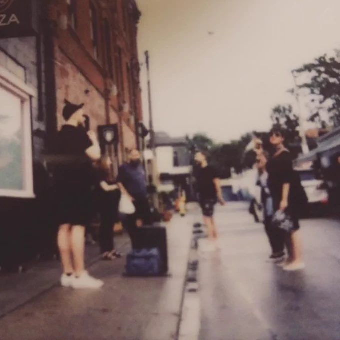 A slightly blurry film photo, designed to look like it was shot in the 1990s, of six members of the Digital 55 team, standing on the street beside a brick building.