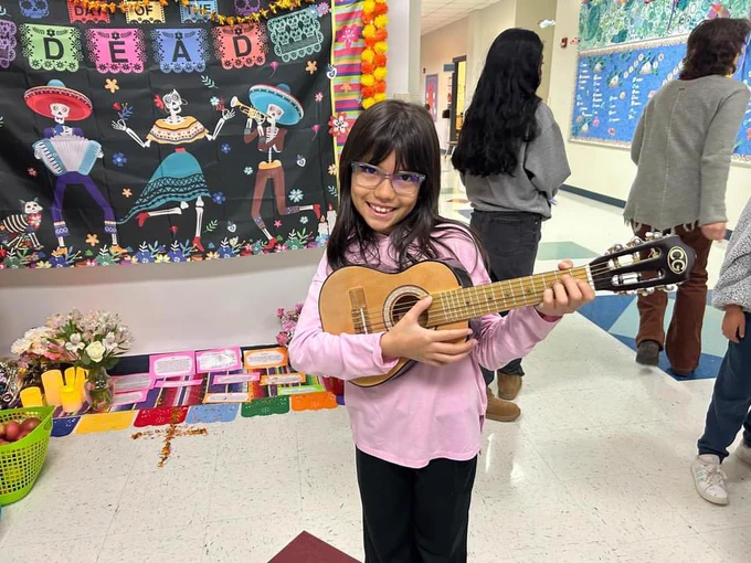 A female student holding a small guitar in front of the ofrenda.