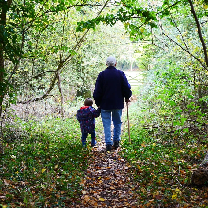 A grandfather walks with his grandson down a forested trail.