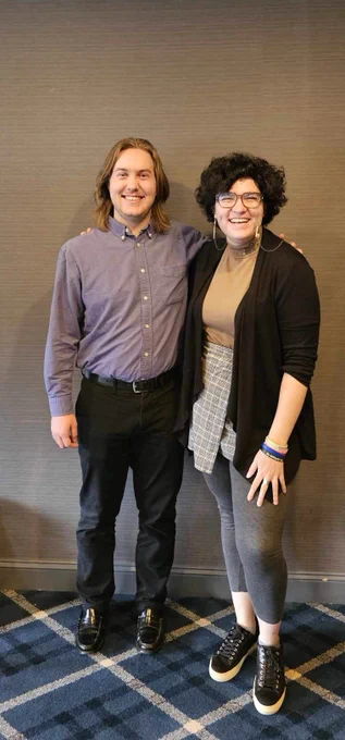 Tarren Sexton (left) and Sarah Carter (right) stand in front of a wall at the conference hotel and smile at the camera. 