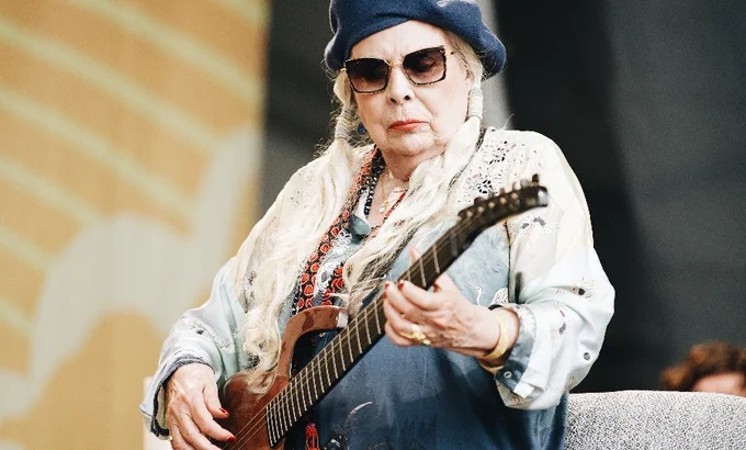 80 years old Joni Mithchell plays guitar wearing white jacket, blue beret and dark sunglasses. 