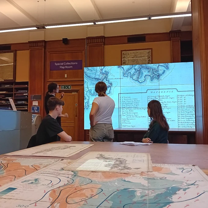 Photo of map room, Main Library. Historic maps are laid out on the large table in the foreground. Four students are examining one of the maps on the large screen in the background.