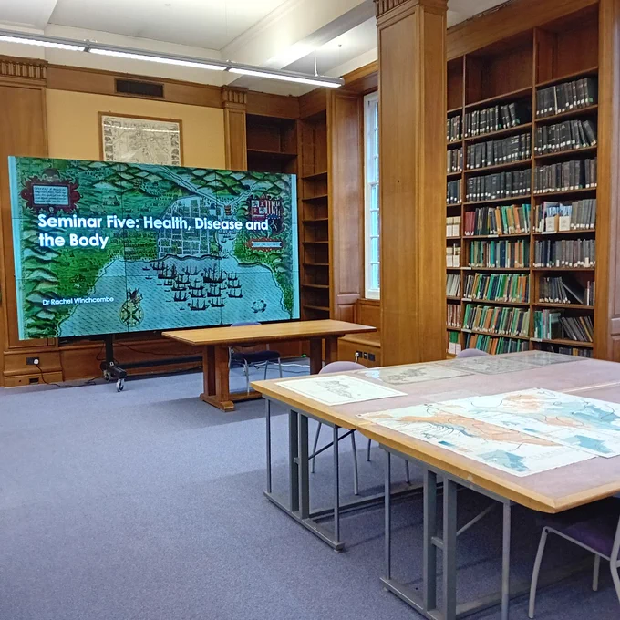 Photo of map room, Main Library. Historic maps are laid out on the large table in the foreground. Shelves of folded maps in the middle distance. A large screen showing a presentation, 'Health, Disease and the Body' by Dr Rachel Winchcombe in the background.