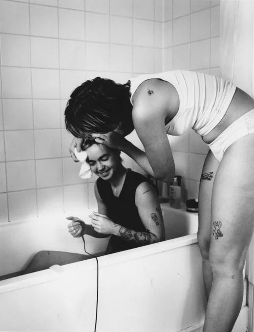 A black and white photo of two people. One is in a bath and the other is washing them with care. 