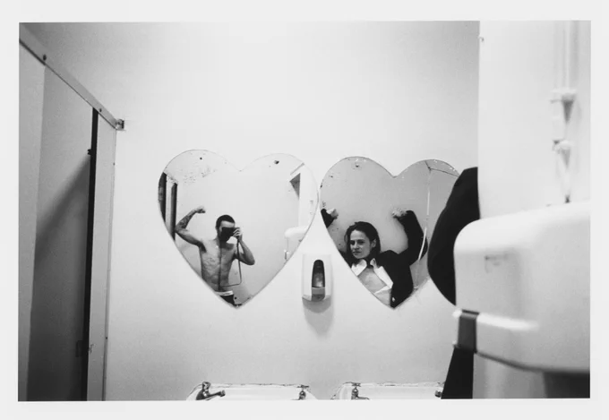 A black and white photo of two people posing for the camera. They are looking into two heart shaped mirrors and doing bicep flex poses