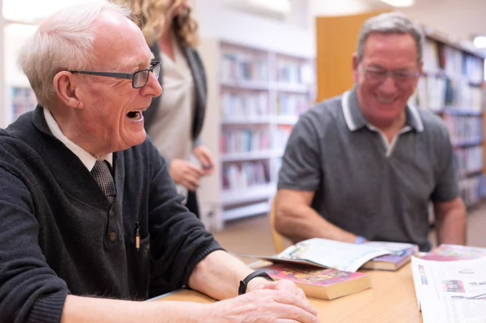 Two older men sat at a table, laughing. There are books on the table top and bookshelves behind them.