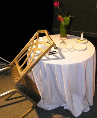a table with a white tablecloth, a plate, a rose in a vase, an inverted glass, and lemon and saltshaker.  A chair is propped by the table.