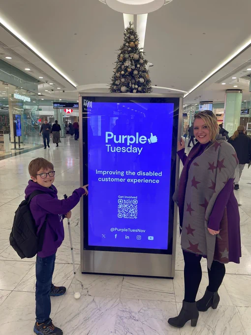 Eastlight resident and Customer Influence Committee Chair, Michelle Baker, with her son, Eli, standing next to a Purple Tuesday sign that reads 'Improving the Disabled Customer Experience' at a London tube station. 