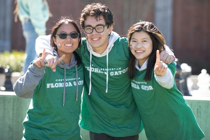 Three students stand together in an embrace wearing green "First Generation Florida Gators" shirts and holding up one index finger to emphasize they are the first in their families to attend college. 