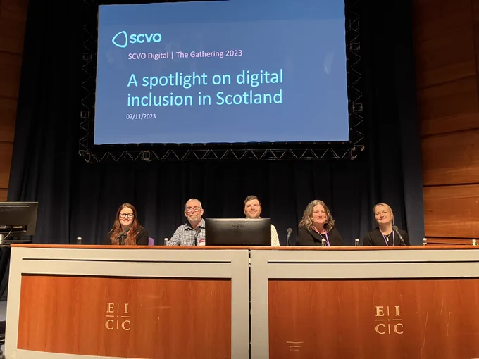 The panel of speakers sits behind a wooden EICC branded desk. On the big screen behind them the slide reads “A spotlight on digital inclusion in Scotland”.