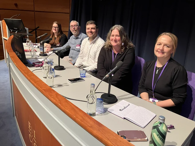 Five speakers on the panel of our digital event sit at a large desk each with a microphone in front of them. They look towards the camera and smile.