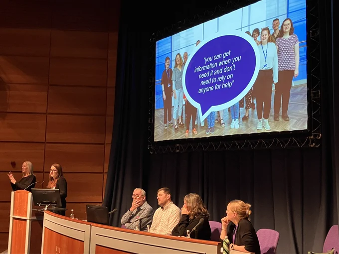 A panel of speakers look on from their seats as one speaker talks from the lectern. Behind the panel is a large screen with a quote on it which reads “you can get information when you need it and don’t need to rely on anyone for help”.