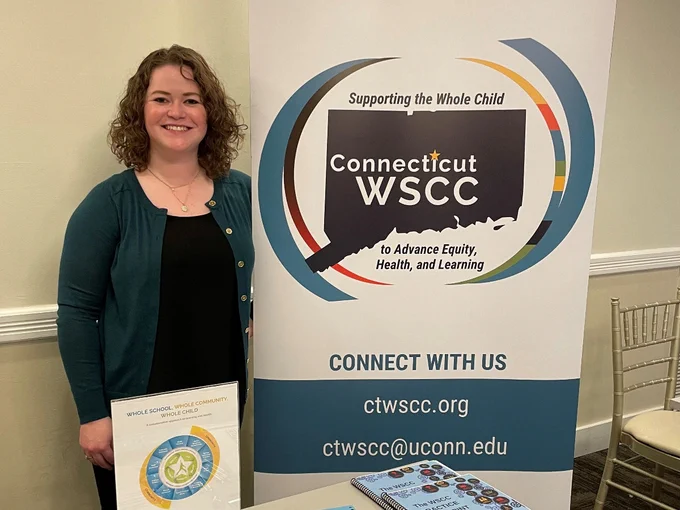 Kate Williamson, dressed in black shirt and dark teal cardigan stands next to banner reading Connecticut WSCC ctwscc.org