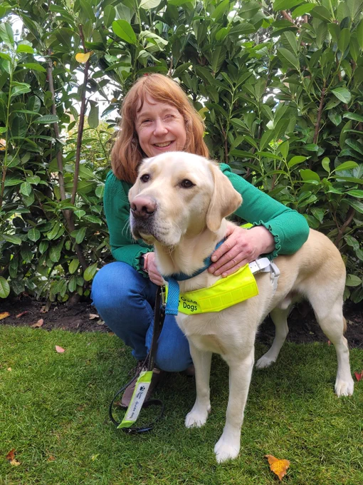 Auto and I in my front garden after we have finished our qualification walk. Otto is a blonde labrador golden retriever and is wearing his guide dog harness and I am wearing a green jumper and jeans. We are sitting together on some grass and I am hugging Otto and smiling.
