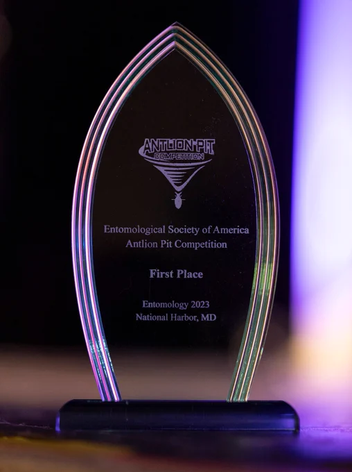 Close-up view of First-Place award trophy for the Antlion Pit Competition. The award is a clear acrylic  peaked oval shape seated on a black pedestal. Engraved on the face is the Antlion Pit Competition logo, followed by text reading “Entomological Society of America | Antlion Pit Competition | First Place | Entomology 2023 | National Harbor, MD”