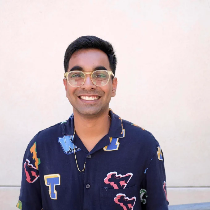 A brown-skinned man wearing light tan, round glasses smiles at the camera. He is wearing a navy button down shirt with capital letters in different fonts decorating it, and a gold chain necklace.