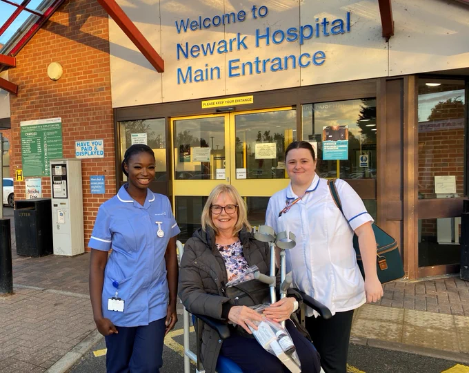 One of the first patients to benefit from a new, state-of-the-art operating theatre prepares to go home. She is pictured outside the main entrance of Newark Hospital. She is in a wheelchair, holding crutches and is accompanied by two uniformed staff from Minster Ward. 