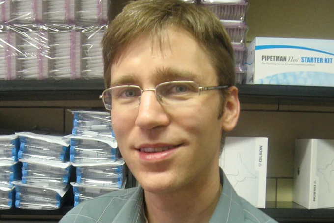 Joel McManus smiles at the camera as he stands in front of biology research supplies.