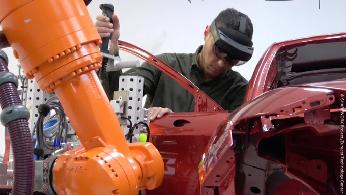 A factory working while performs tasks for the automation of transportation of automotive pieces and operators promoting the idea of collaborative robots.