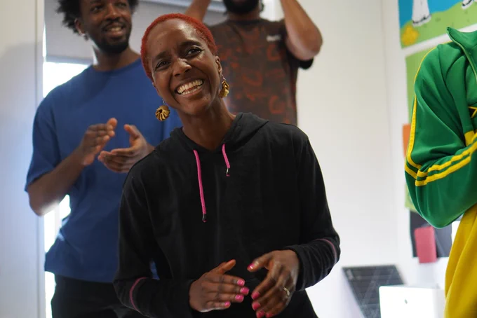A person with very short red hair, gold hoops and a black hoodie, laughing and smiles in yard. Behind them people are moving and clapping hands together.