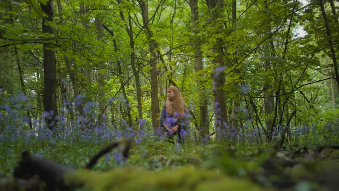 Director Ella Glendining stands in the centre on the frame; she is a white woman in her twenties with long blonde hair. Lush and vibrant forest takes up the frame, with bluebells in the foreground. Ella looks over her shoulder, ethereal and slightly blurred.