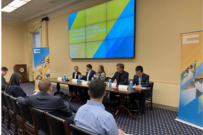A panel of five University of California researchers addresses an audience at a briefing. They are flanked by two pop up banners and a large video screen promoting UC. 