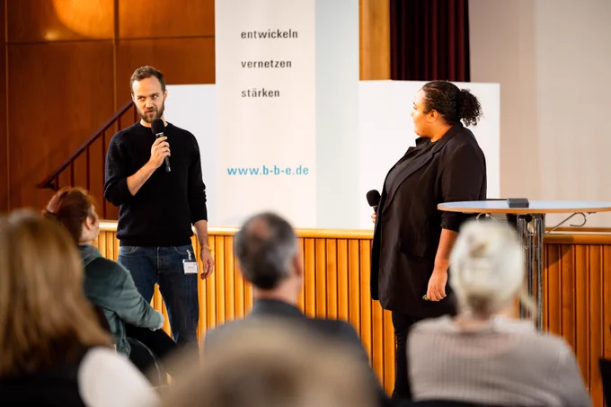 Auf dem Podium stehen zwei Personen, links ein junger Mann in Jeans und schwarzem Rollkragenpulli, mit Mikro in der Hand. Rechts eine junge Frau in schwarzem Blazer und mit Mikro in der Hand