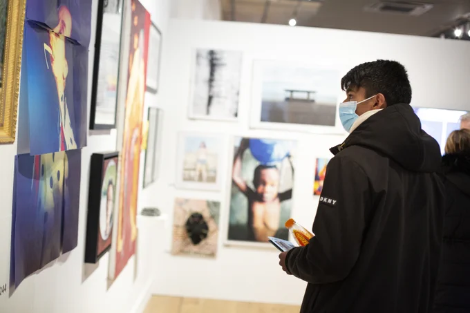 Photo © Mark Savage / Bolton School. A teenage boy stands in a black coat with a blue mask on his face, looking to the wall in front of him which has a variety of art pieces hung on. 