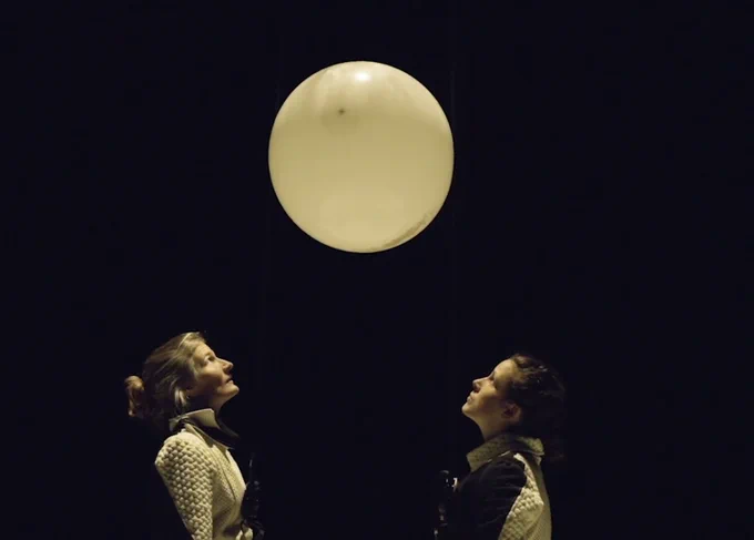 Black background, two brunette females, looking up at a white ballon above their heads. 