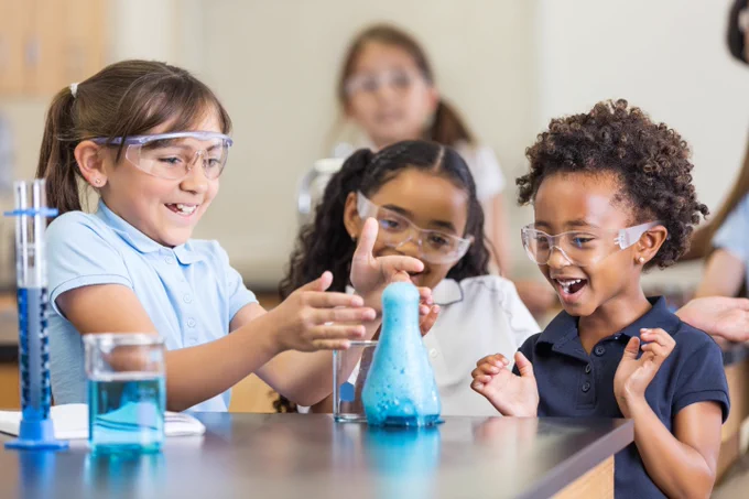 Three smiling kids with protective eye  equipment while standing around a beaker overflowing with blue liquid