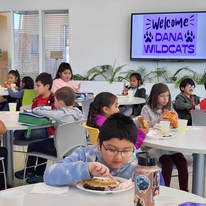 Over 10 students sitting at tables eating pizza with a sign behind them that says "Welcome Dana Wildcats."