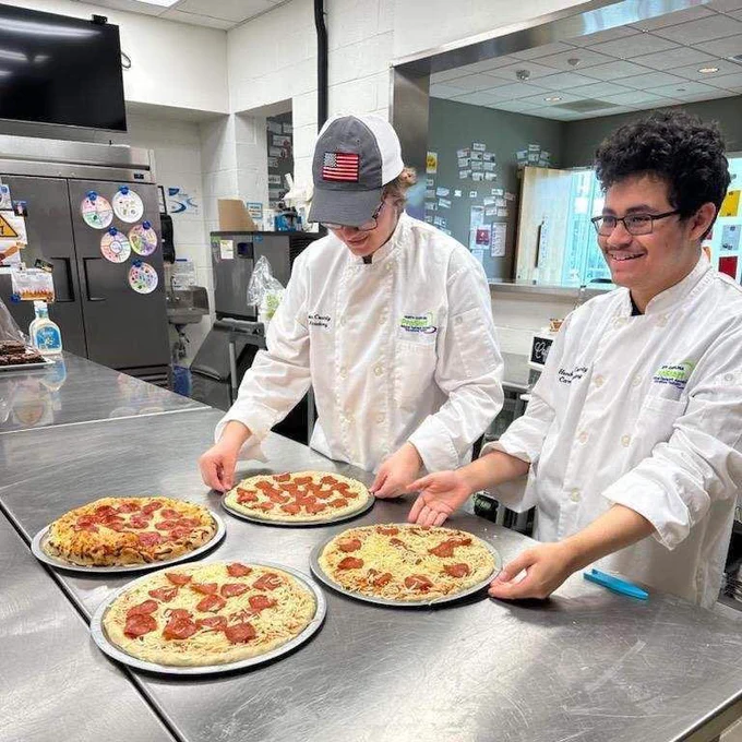 Two culinary students standing in the kitchen with four pepperoni pizzas.