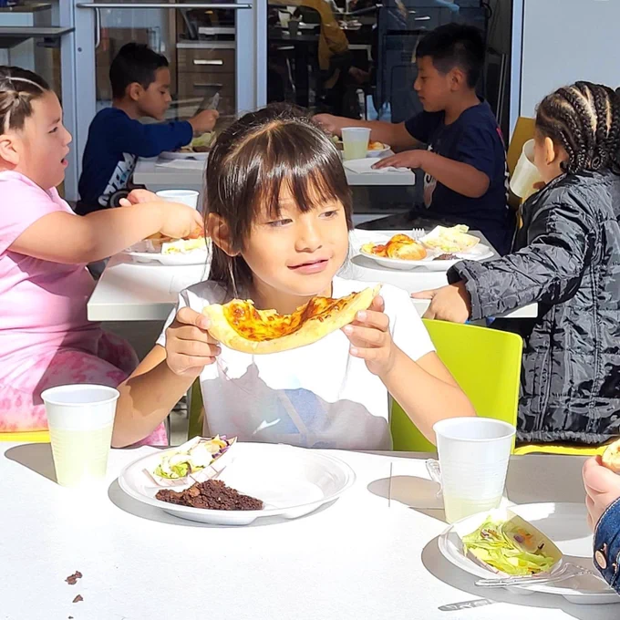 A student holding a big piece of pizza with four students eating behind her.