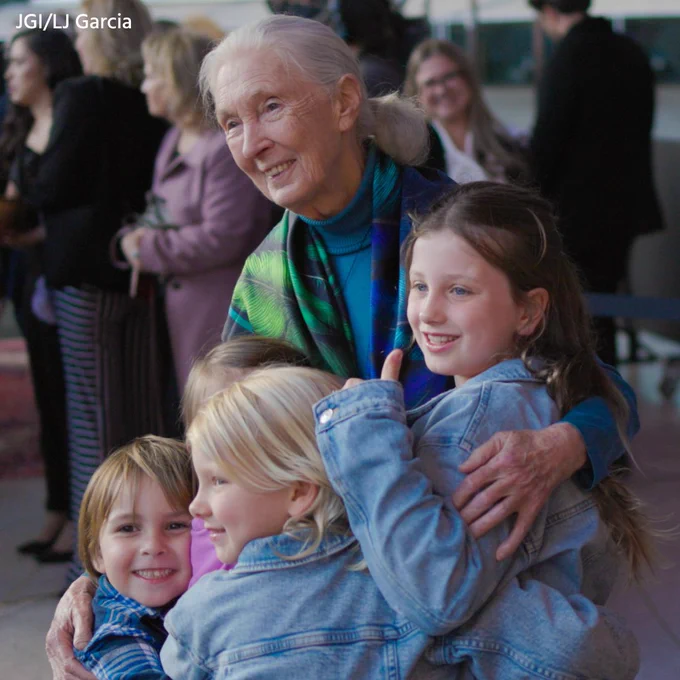 Photo of a pale, elderly woman with white hair pulled into a ponytail (Dr. Jane Goodall) smiling and hugging four smiling children of difference ages and appearances. Photo credit: JGI/LJ Garcia.