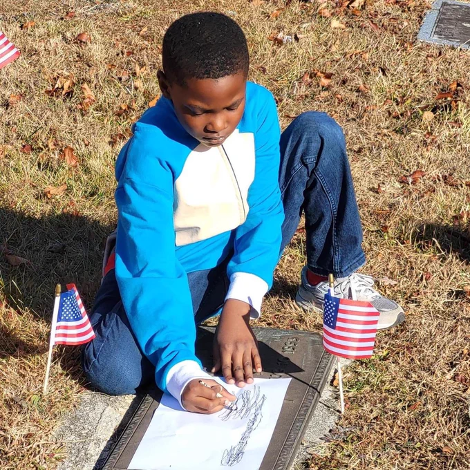 A student with a crayon and paper trying to trace a veterans name at the grave.