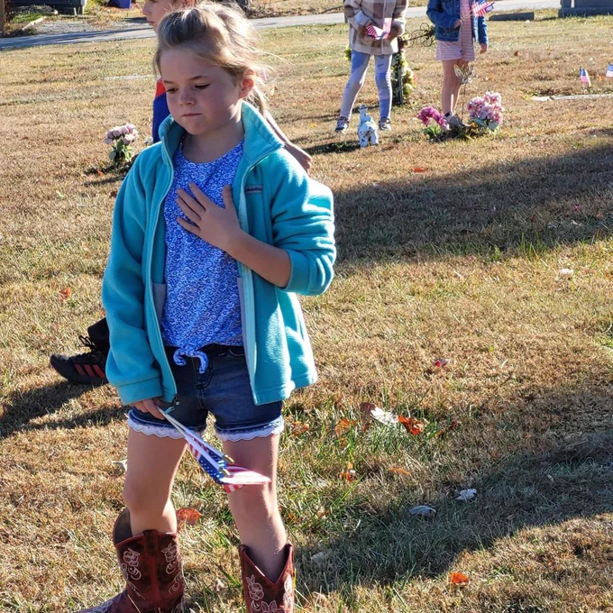A student holding a hand over their heart while holding an american flag at the grave.