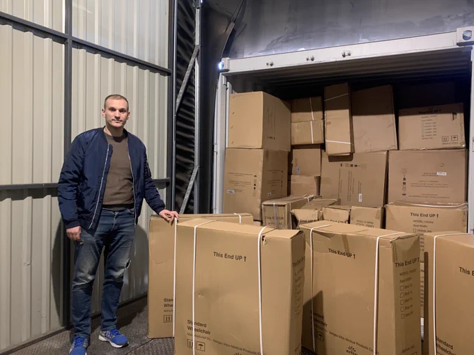 A man in jeans and a blue jacket stands in front of a container full of wheelchair boxes.